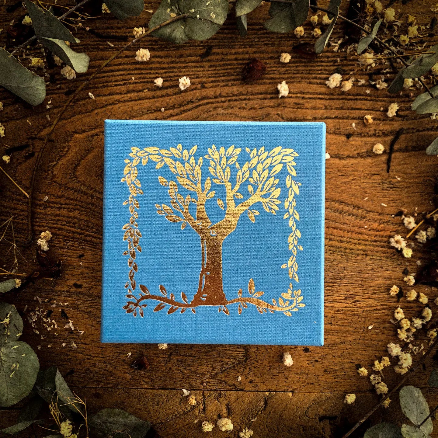 A bright blue Spiramirabilis box painted with a gold tree, among eucalyptus